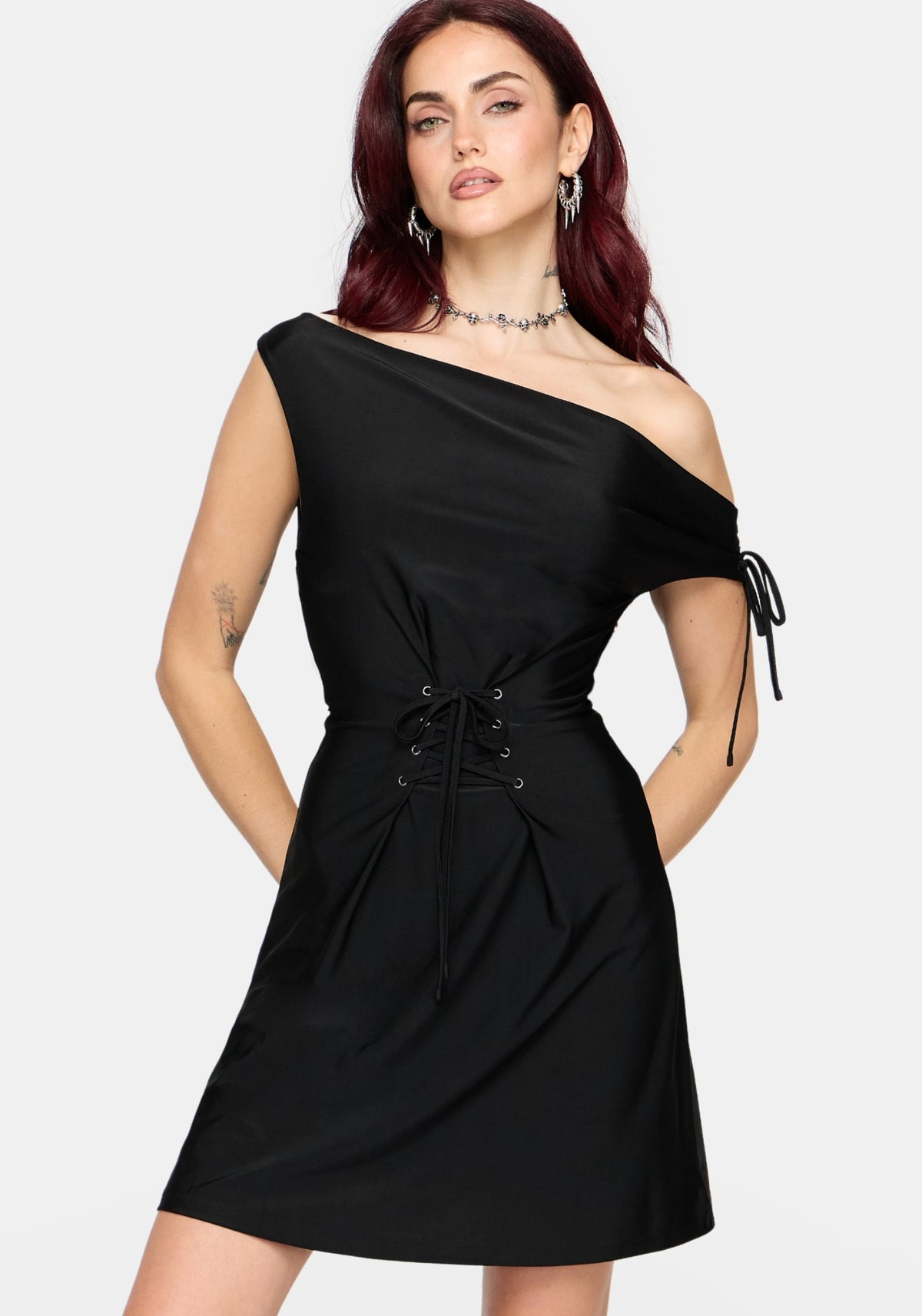 A woman modeling a black one-shoulder dress with a short sleeve, side tie detail, and lace-up waist against a light gray background.
