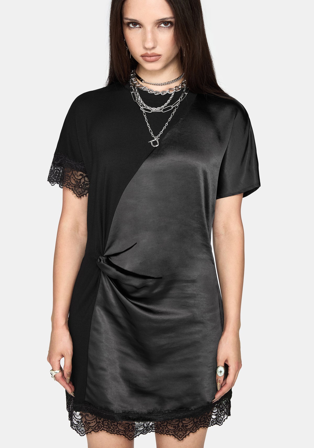 A woman modeling a black satin-like short-sleeve dress with lace trim at the sleeve cuffs and hem, layered with silver chain necklaces, against a light gray studio background.