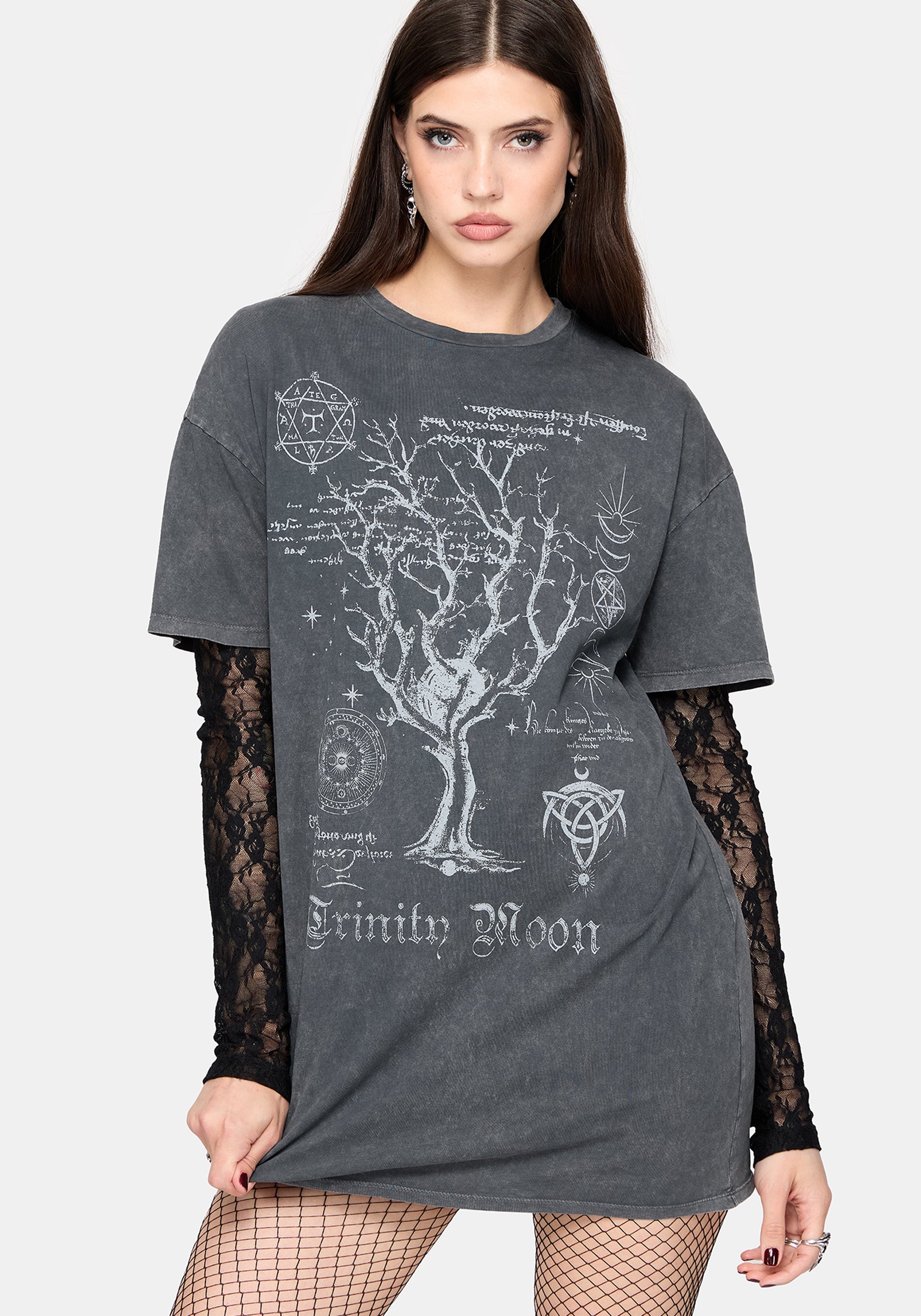 A woman modeling a dark gray acid-wash t-shirt with a white gothic tree and occult graphic print, layered over a black lace long-sleeve top and fishnet tights against a light gray studio background.