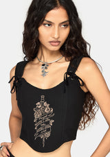 A woman wearing a black sleeveless corset top with side tie details and a beige graphic print on the front, photographed against a light gray studio background.